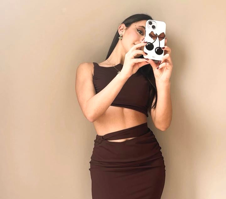 A woman in a crop top and high-waisted skirt taking a mirror selfie, holding a phone with a cherry-shaped case adorned with brown bows and cherries.