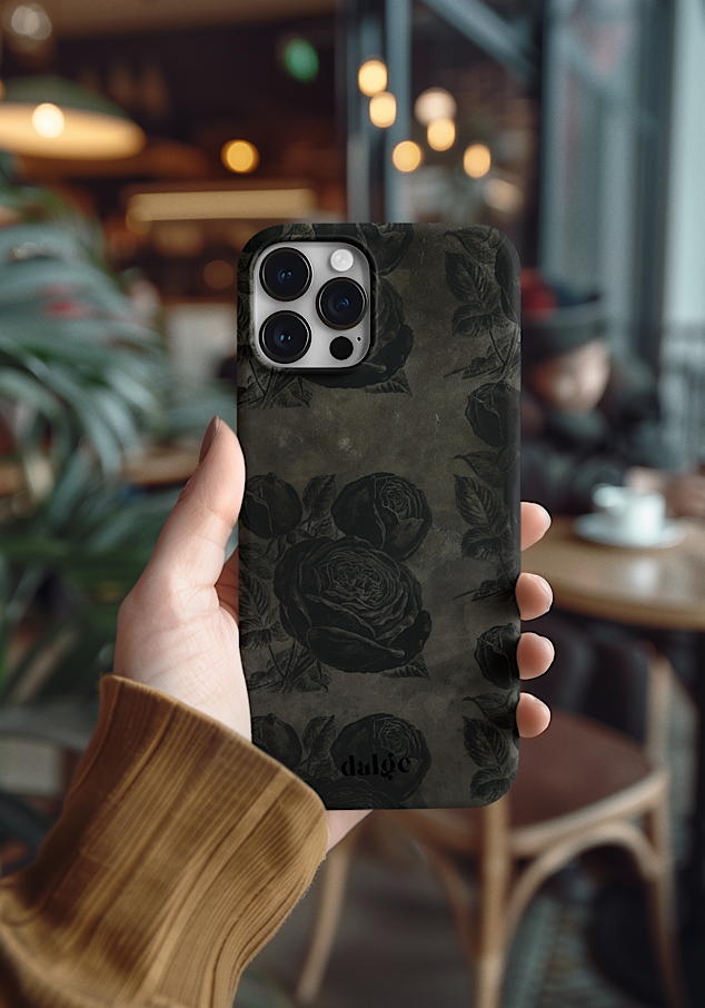 A person holding an iPhone with dark floral academia phone case, featuring the brand name 'dalge' at the bottom, against a blurry coffee shop background