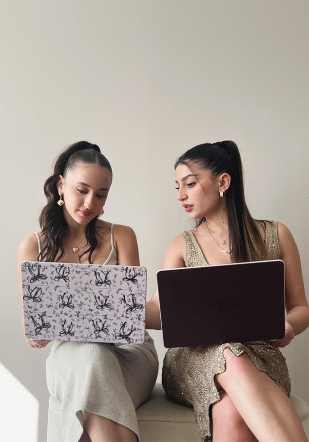 Two women seated with laptops—one featuring a black and pink bow design cover, the other in solid burgundy.