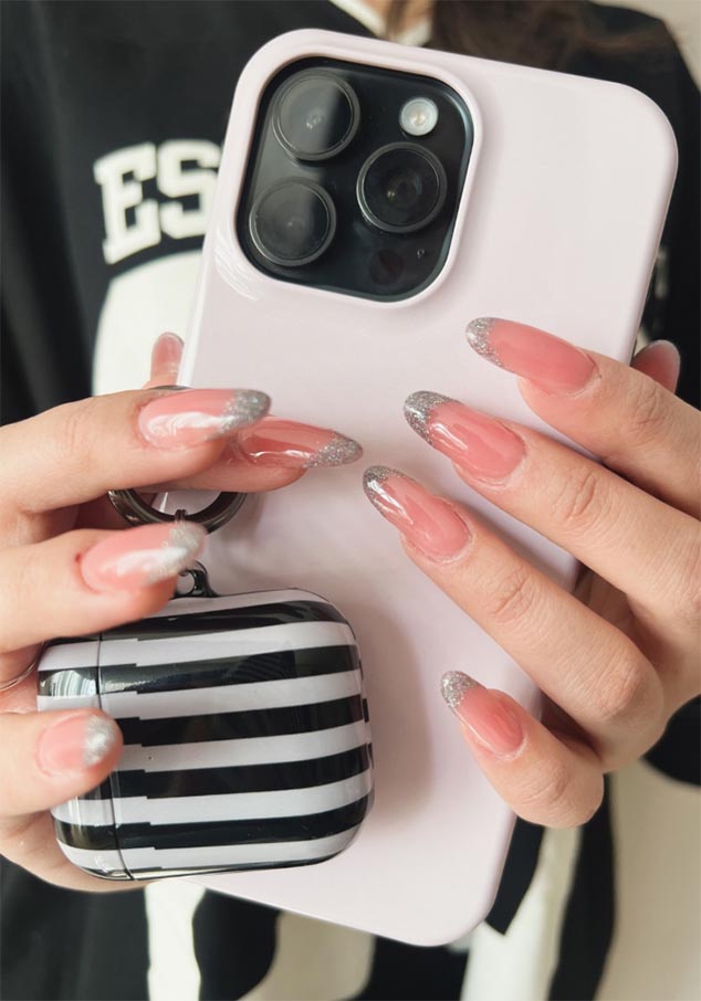Hand holding a pink smartphone with a triple-lens camera, glittery silver nail tips, and a black-and-grey striped AirPods case.