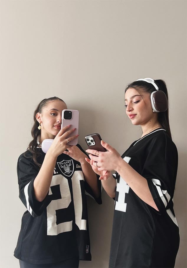 Two women holding iPhones with pink and burgundy cases, each wearing glossy burgundy and pink AirPods Max cases respectively.