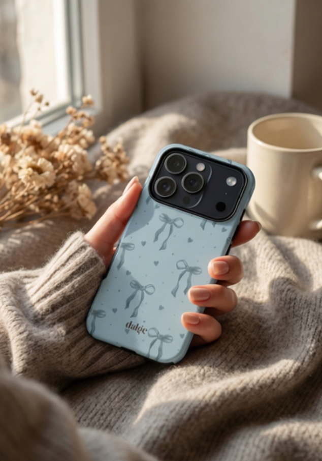 Person holding a phone with a blue case featuring bow designs, sitting on a soft surface with a cup and dried flowers in the background.